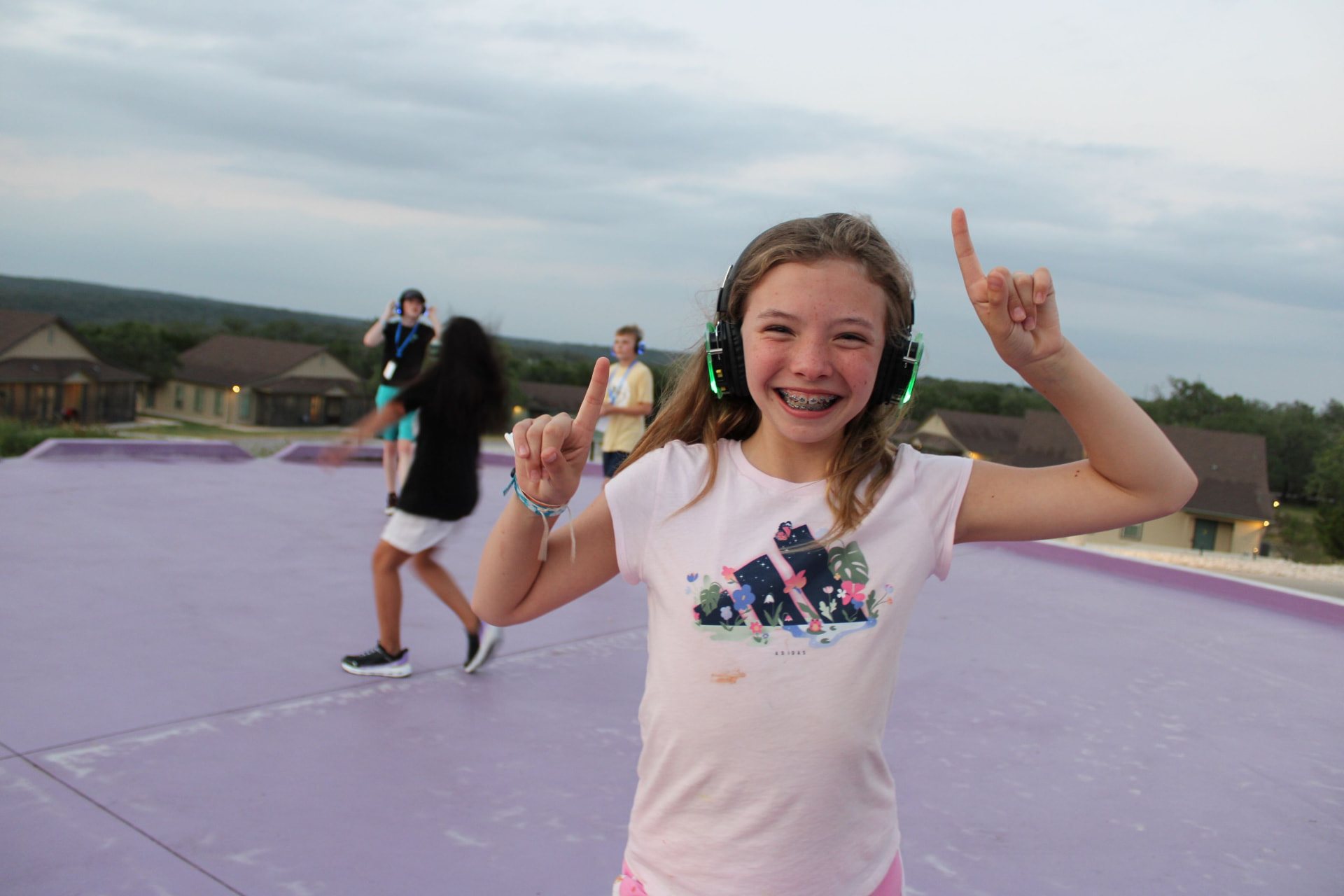 A camper in headphones throws her hands up dancing on the camp court at dusk.