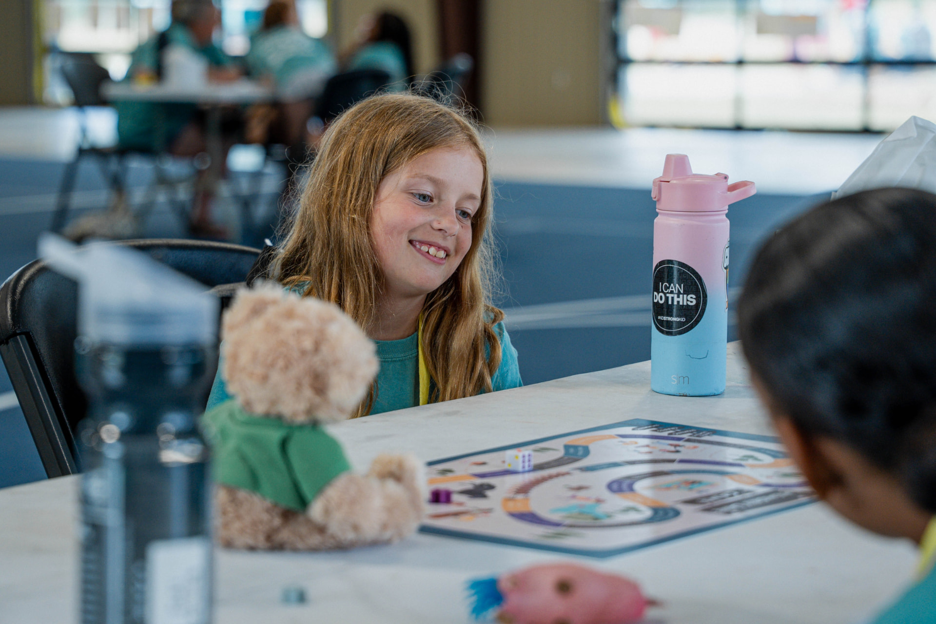 A smiling girl plays the Camp Agape Memory Lane Game at a table with her teddy bear beside her.