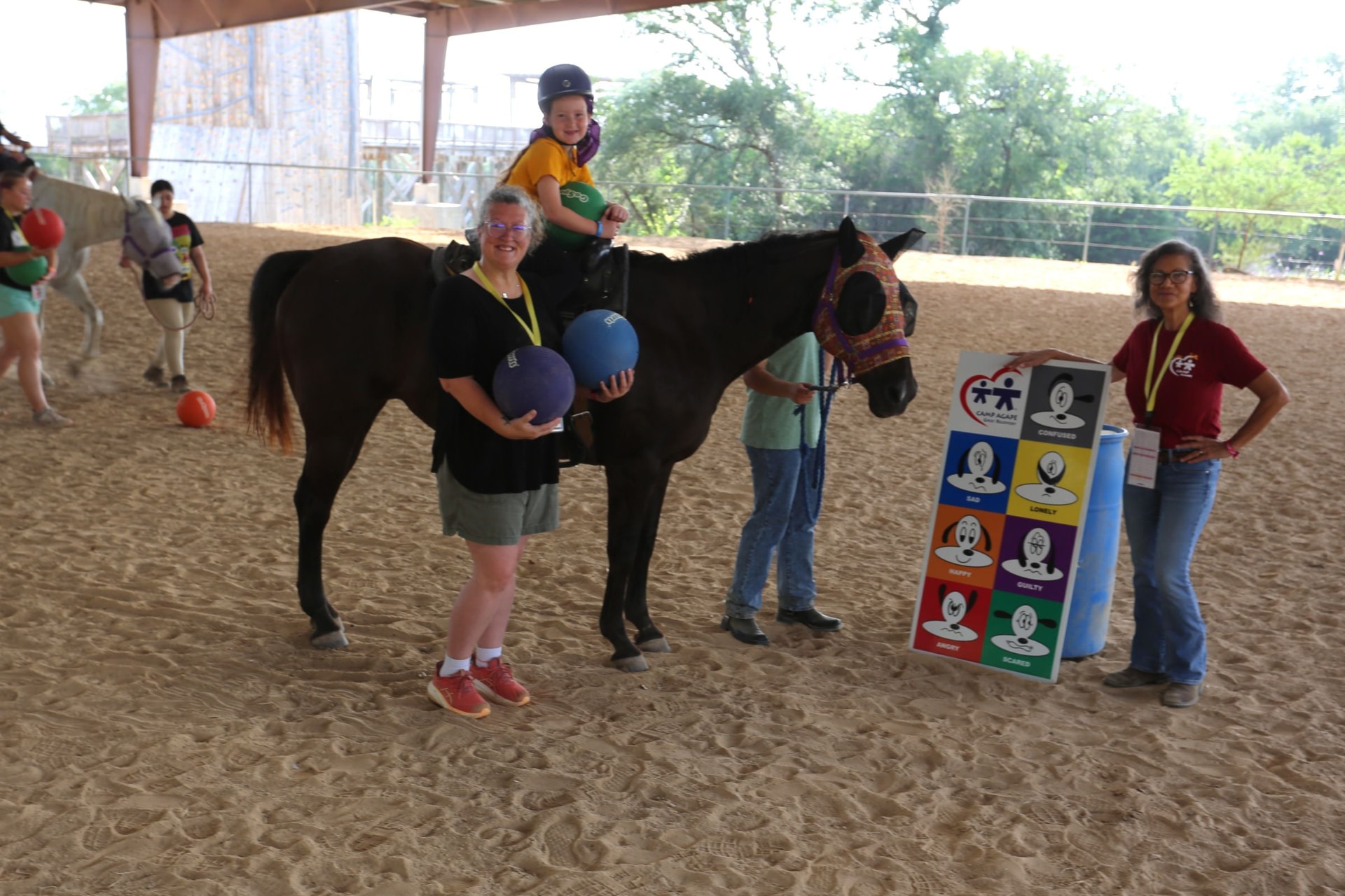 Volunteers and a camper on horseback beside the Camp Agape Emotion Board in the riding arena.