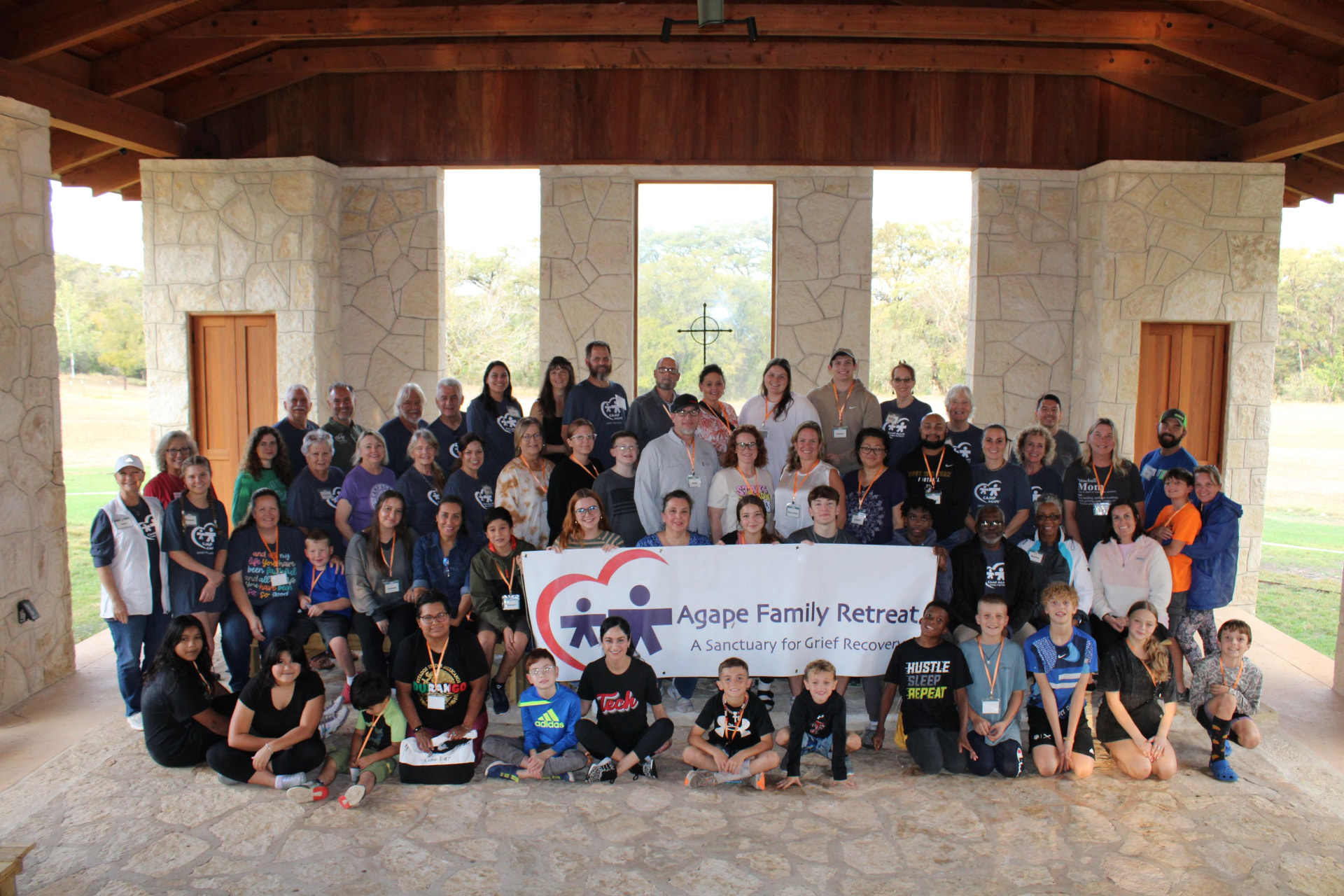 A large group of adults and children pose together under a pavilion holding an Agape Family Retreat banner.