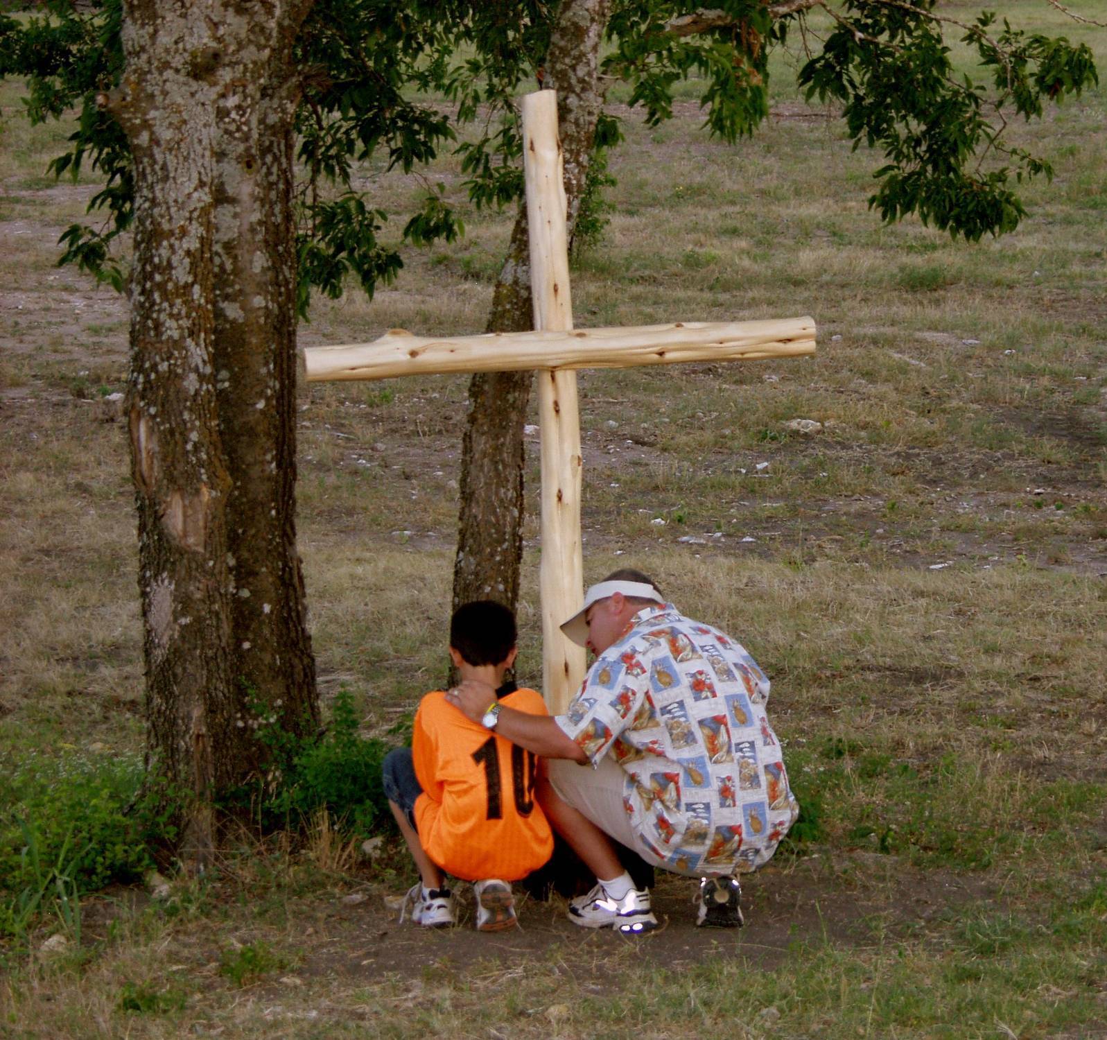 A volunteer kneels beside a camper in an orange shirt at a wooden cross during an outdoor Camp Agape moment.